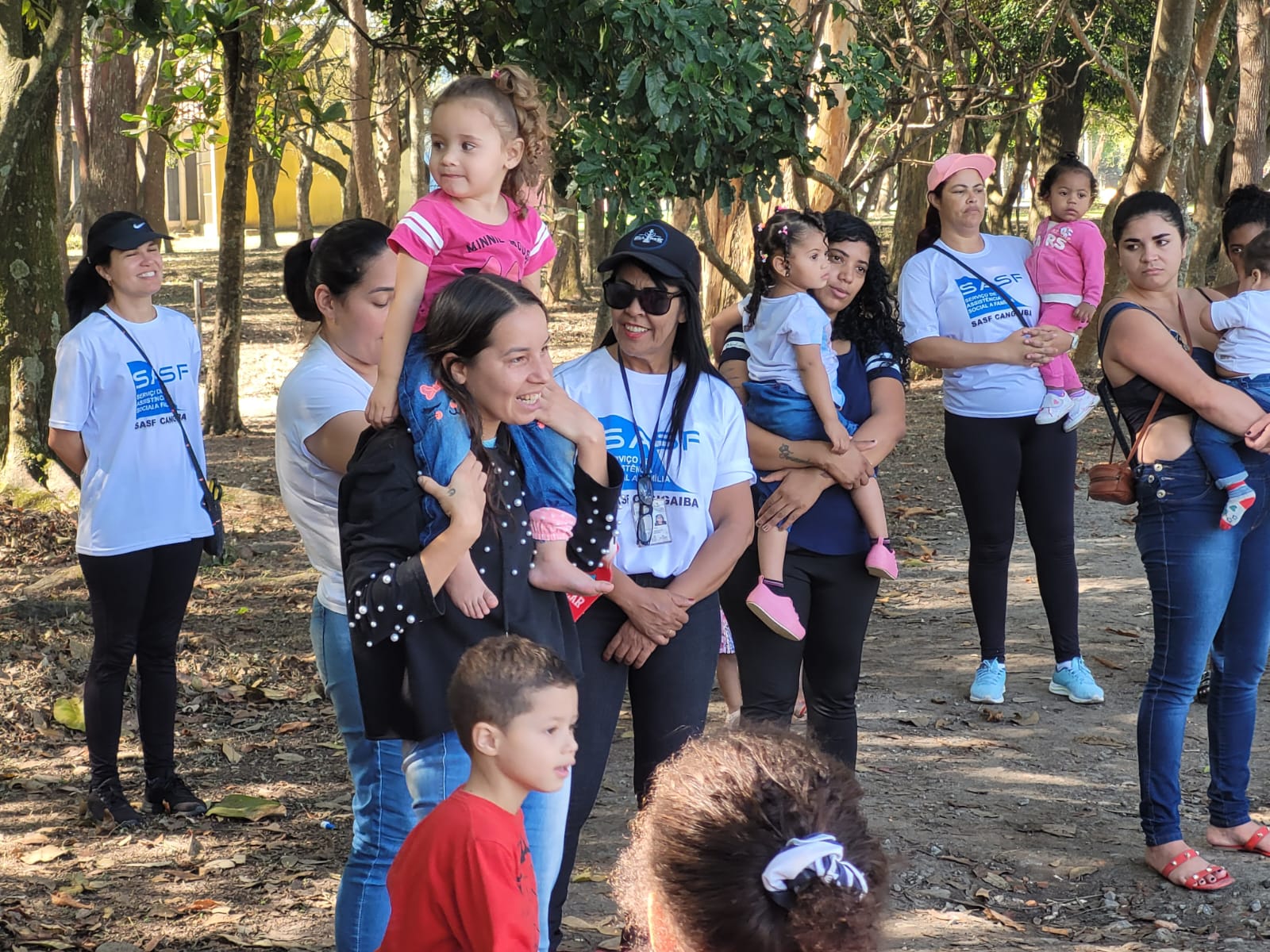 Há várias pessoas em frente a algumas árvores, funcionários da SASF vestidos de branco com a logo azul da instituição, junto com mães e suas crianças, uma delas de preto com a filha vestida de rosa em seus ombros, outra de azul com a filha vestida de cinza no ombro. Na frente tem um menino vestido de vermelho.